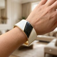 لقطة مقربة لامرأة ترتدي في معصمها سواراً ذهبياً عريضاً ومكتنزاً (Cuff Bracelet) يتميز بتصميم هندسي مقسم إلى مثلثات من المينا البيضاء اللامعة، يتوسطها شكل معين من المينا السوداء ذات الملمس البارز، على خلفية داخلية ضبابية.