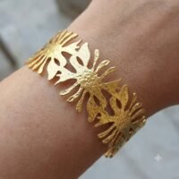 لقطة واقعية لسوار ذهبي عريض ومفرغ (Cuff Bracelet) بنقوش متكررة تشبه زهور اللوتس الفرعونية أو أشعة الشمس، مغلف داخل كيس بلاستيكي شفاف يظهر عليه جزء من كلمة "Stainless steel".